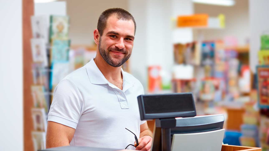 Homem sorrindo atrás de um caixa de loja, ao lado de um sistema de ponto de venda (PDV), em um ambiente de varejo com prateleiras de produtos ao fundo.