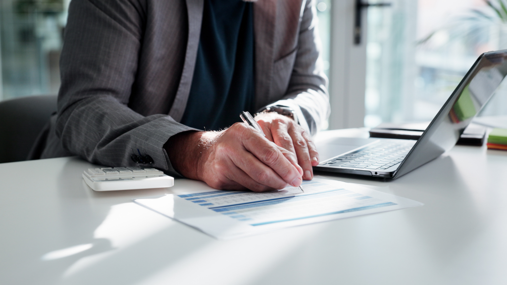Homem de terno sentado à mesa de escritório, utilizando um notebook enquanto preenche um documento financeiro com uma caneta; ao lado, há uma calculadora. A cena transmite análise de dados, controle financeiro e planejamento empresarial.