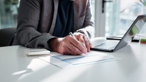 Homem de terno sentado à mesa de escritório, utilizando um notebook enquanto preenche um documento financeiro com uma caneta; ao lado, há uma calculadora. A cena transmite análise de dados, controle financeiro e planejamento empresarial.