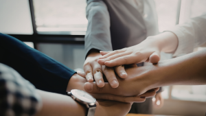 Grupo de pessoas em ambiente corporativo com as mãos empilhadas no centro, simbolizando união, colaboração e trabalho em equipe.